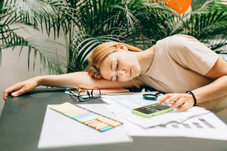 Woman lying on table with her glasses off trying to fill out her tax forms using her phone calculator