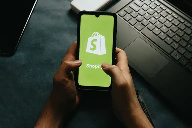 Hands Holds A Cell Phone Showing The Shopify Logo Photo