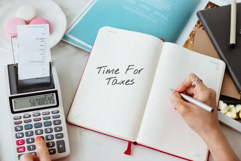 Close-up on a woman's hands while working on a calculator with one and holding a pen in the other as it hovers over a notebook with the words “Time for Taxes” written in it