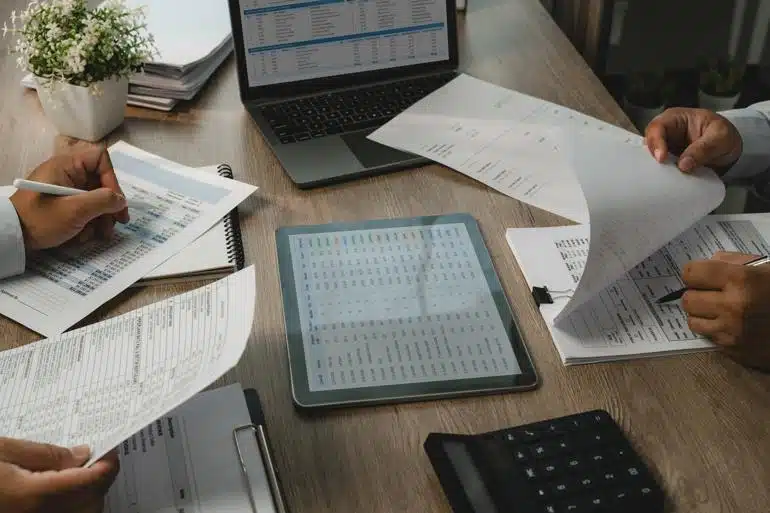 Two sets of hands checking paperwork at a desk with a calculator, tablet, and laptop on it